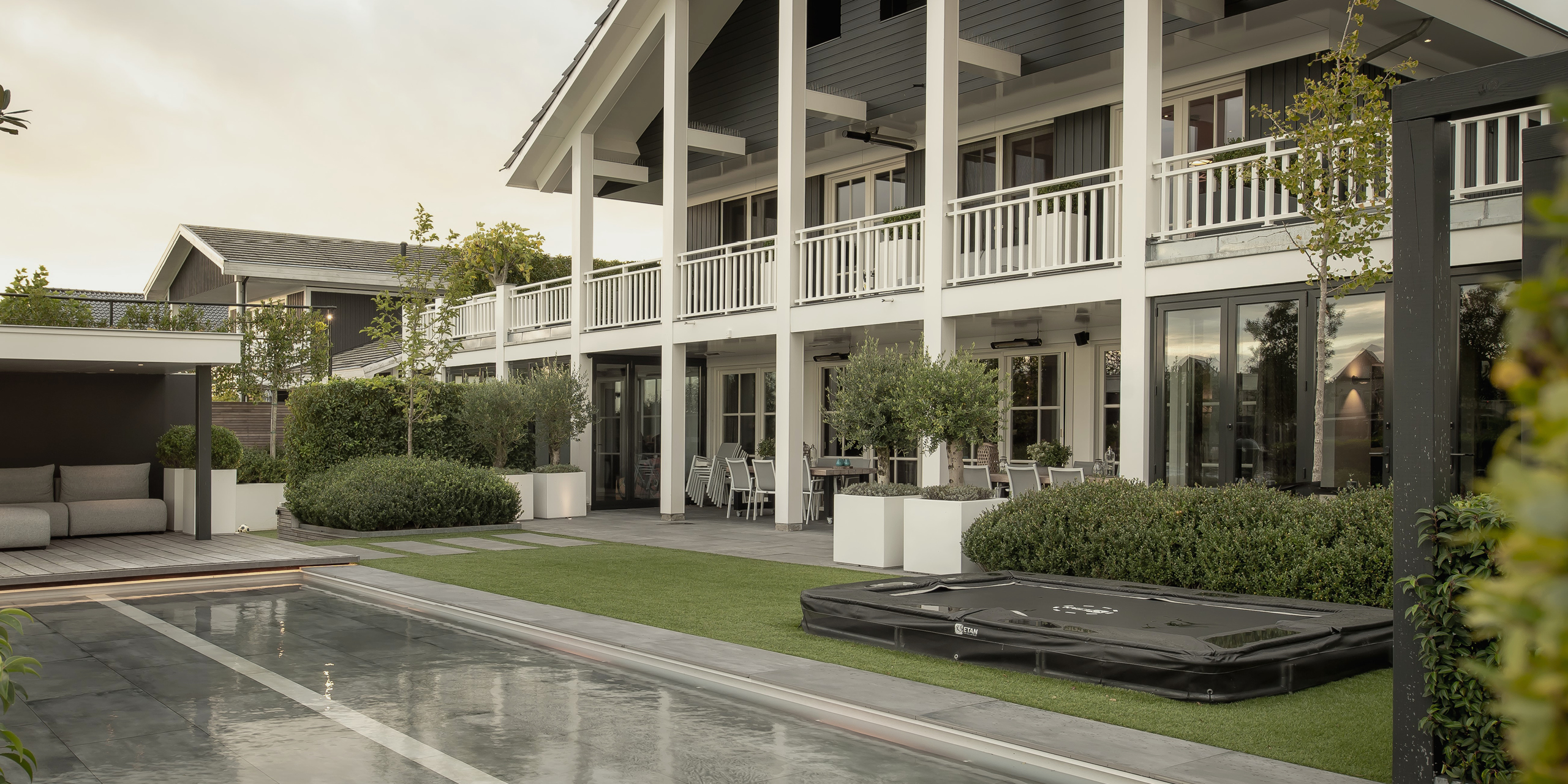Buytengewoon wellnesstuin in Leeuwarden met veranda, loungehoek, vlonder, liguster, olijfbomen, terras, zwembad en trampoline.