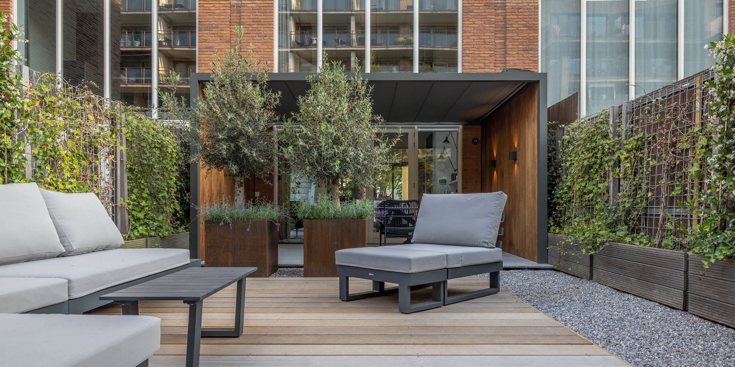 Amsterdamse Buytengewoon stadstuinen met loungehoek, vlonder, bloembakken en olijfbomen.